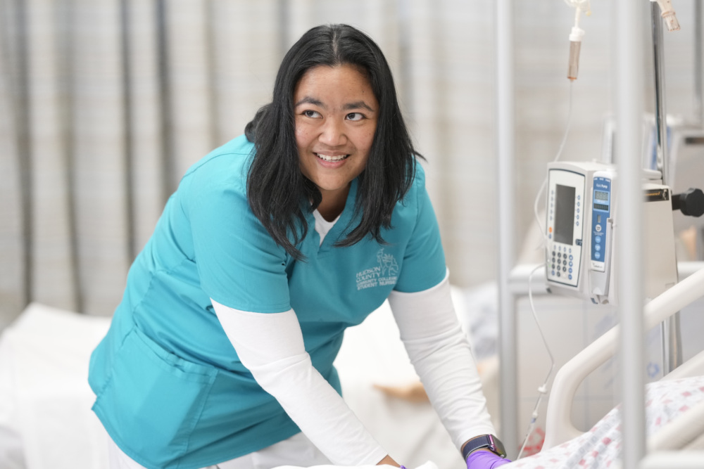 A nurse in blue scrubs smiles while adjusting equipment next to a hospital bed in a clinical setting.